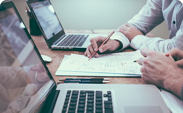 Close-up view of two people's hands reviewing a handwritten financial document or plan on a desk, with two laptops open in the foreground and background.