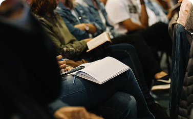 Close-up of audience members sitting in rows; one person is resting an open blank notebook on their knee, holding a pen, while another person nearby holds an open book.