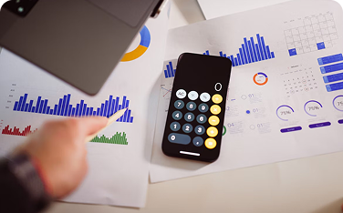 Overhead shot of a hand pointing a pencil at multiple printouts of bar and pie charts. A black smartphone showing a calculator app is placed in the center of the documents.