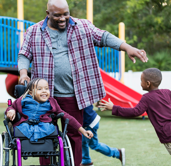 A happy father smiles while pushing his young daughter in a wheelchair (who is also smiling broadly), as her brother runs toward them with arms outstretched for a hug in a colorful playground.