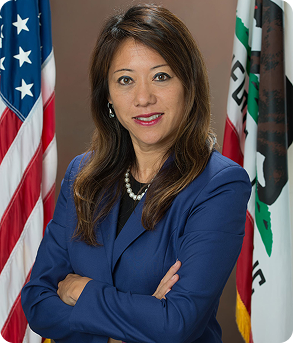 Formal portrait of Fiona Ma, California State Treasurer, wearing a blue suit and pearls, standing with her arms crossed between the US and California flags.