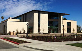 Exterior view of a modern, beige and glass institutional building with a flat roof, surrounded by concrete walkways and landscaping under a bright blue sky.